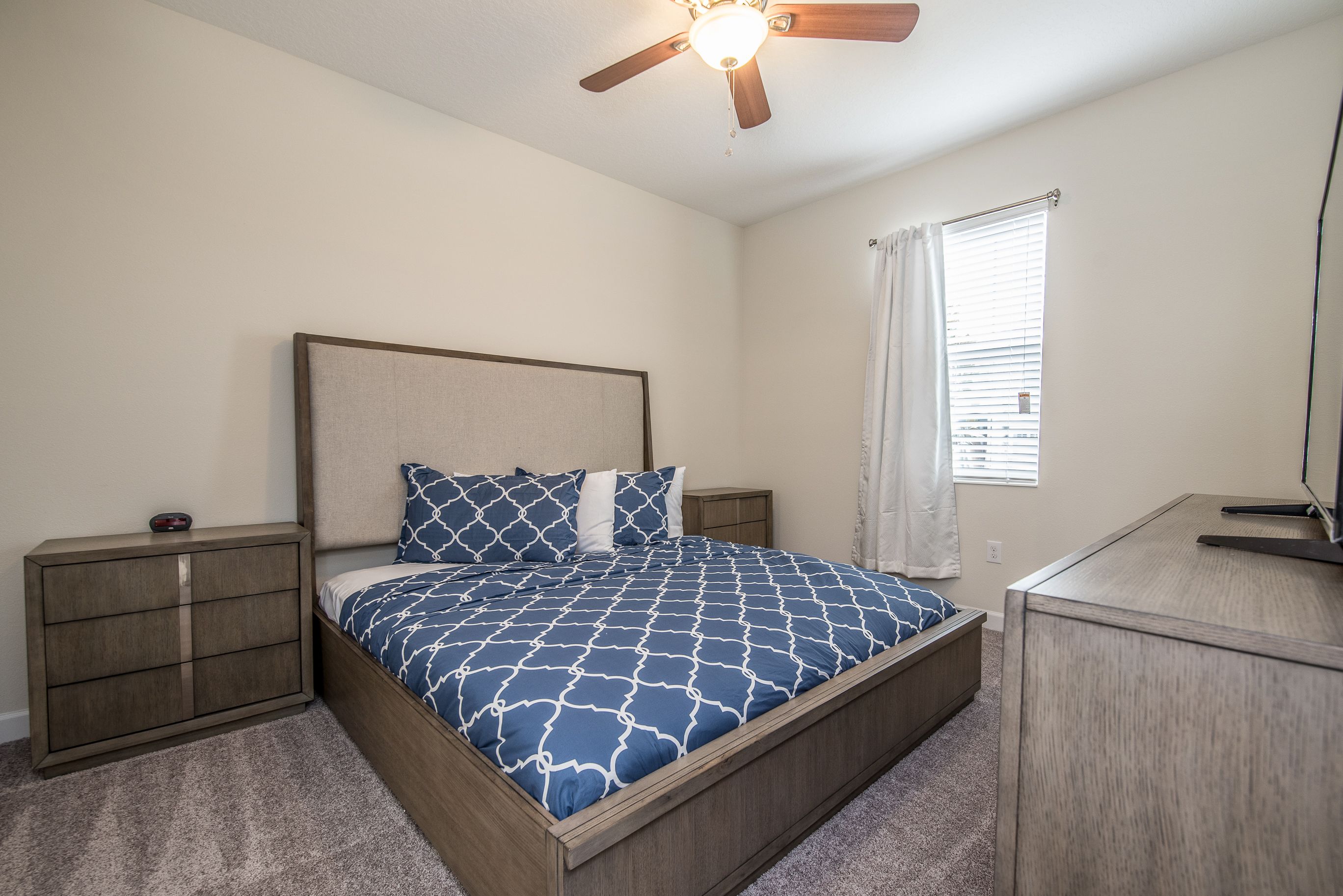 A bedroom with a bed, nightstands, a desk, a ceiling fan, and a window with blinds.