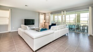 A spacious living room with a white sectional sofa, a flat-screen TV, and a dining area with glass doors leading to a balcony.