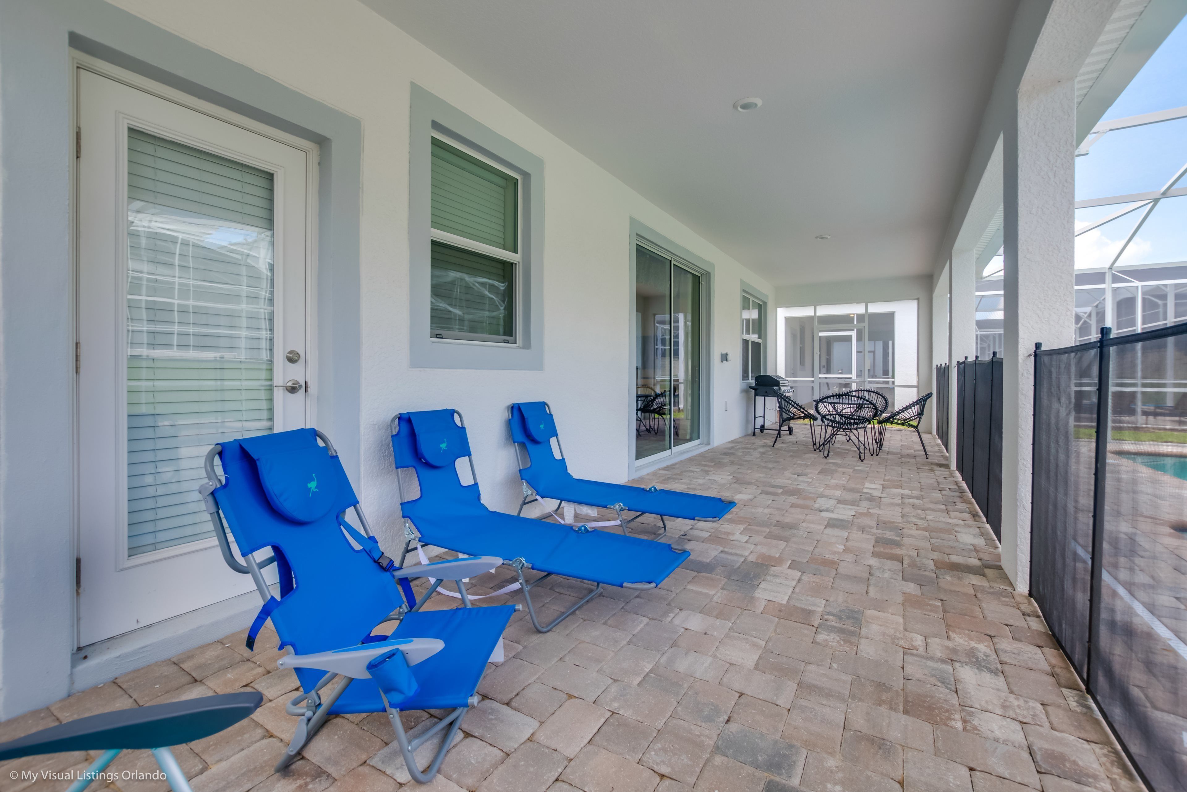 A covered outdoor area with blue lounge chairs and a table, a glass door, and a view of a pool.