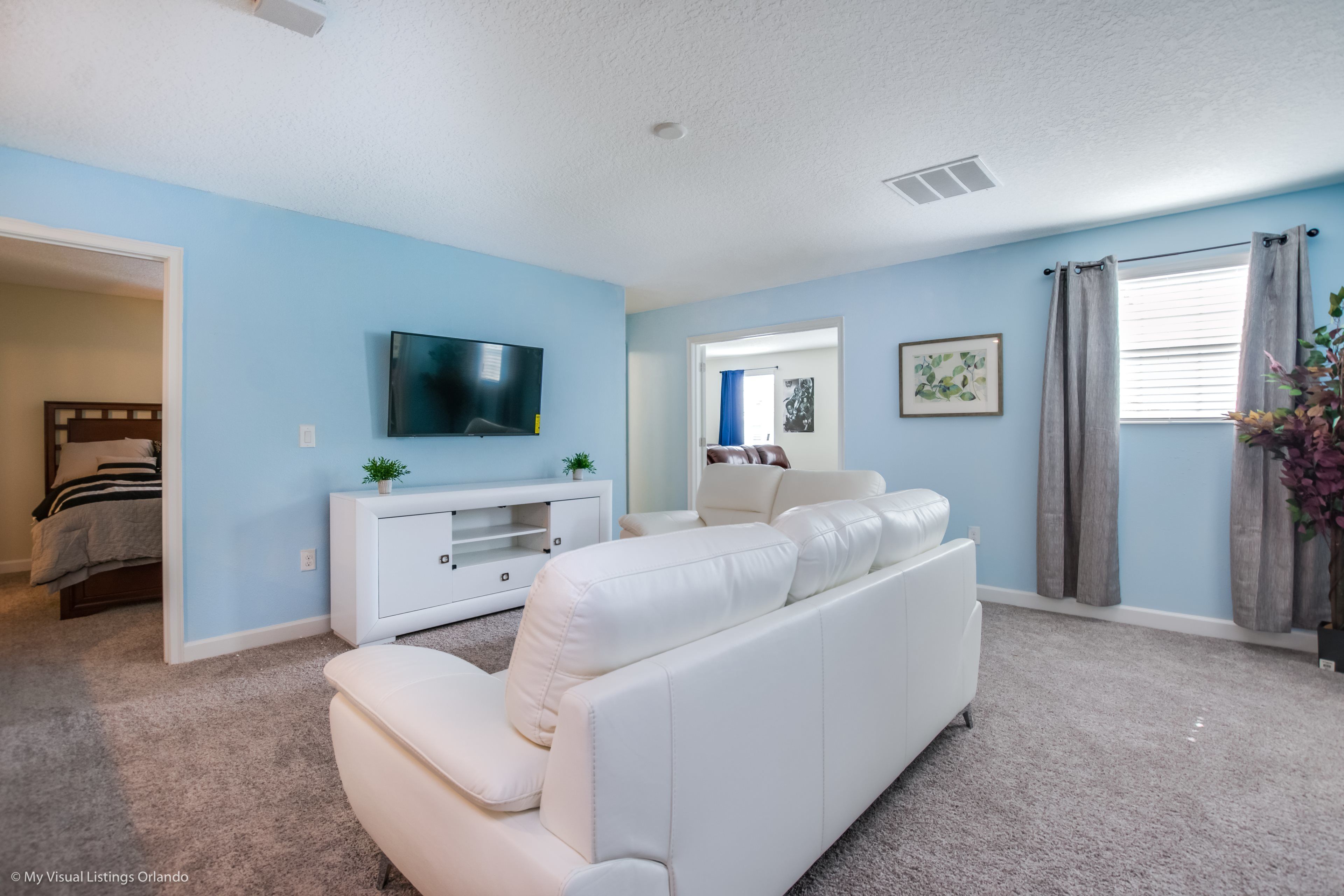 A living room with a white sofa, a TV mounted on the wall, a small cabinet, and a window with curtains.