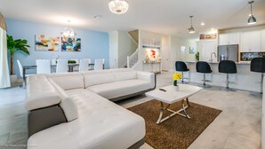 Un salon moderne avec un canapé d’angle, un coin repas avec une table et des chaises blanches, et une cuisine avec des meubles blancs et des tabourets de bar.