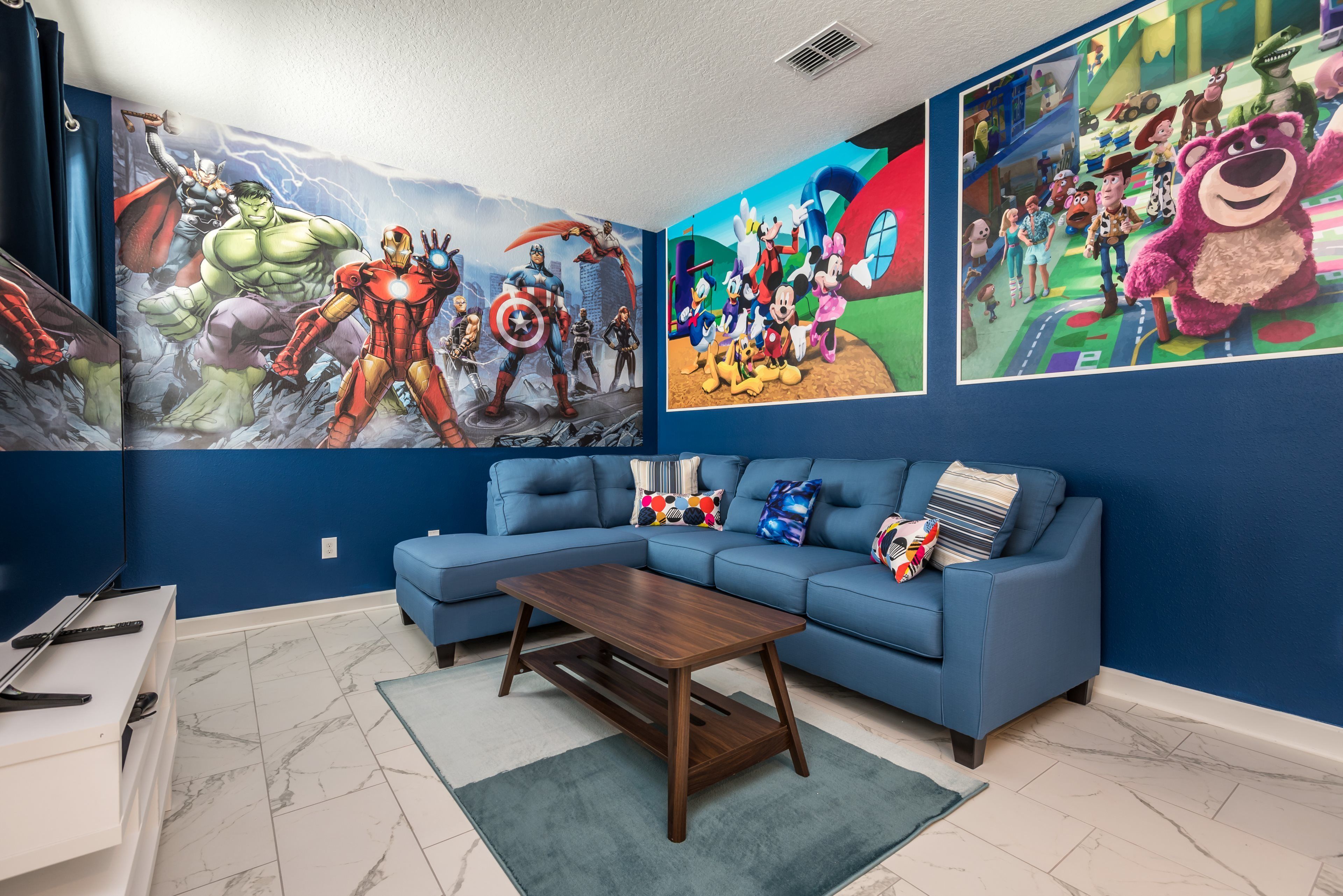 A living room with a blue sofa, a coffee table, and a wall mural featuring various animated characters.