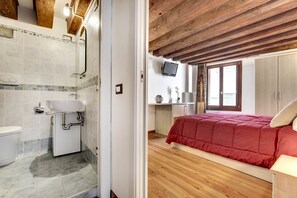 A bathroom with a sink, toilet, and shower. The bedroom has a bed with a red blanket, wooden flooring, and a TV on the wall.
