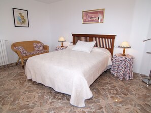 Un dormitorio ordenado con una cama grande, dos mesitas de noche con lámparas, una silla de mimbre y dos cuadros en la pared.