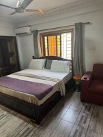 Un dormitorio con una cama, ventilador de techo, equipo de aire acondicionado, ventana con cortinas, una silla roja y un taburete pequeño.