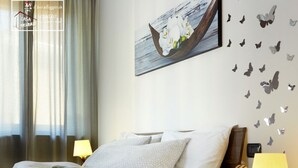 Un lit bien fait, recouvert d’une couverture à motifs, une table de chevet avec une lampe et des livres, un tableau représentant un bateau accroché au mur, et des autocollants décoratifs en forme de papillons.