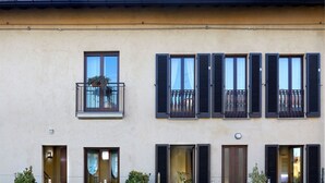 Un bâtiment doté de nombreuses fenêtres, dont certaines sont équipées de volets, et de balcons avec des garde-corps. On y trouve des plantes en pot et une table à l’extérieur.
