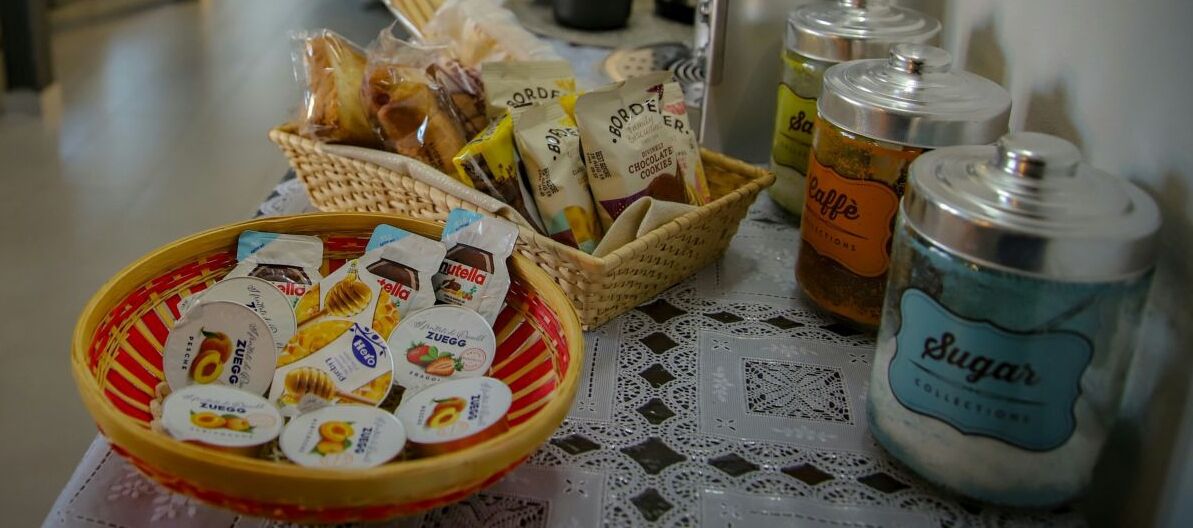 Un tavolo con un cesto di snack confezionati, un barattolo con l'etichetta 'Sugar' e una ciotola di biscotti.