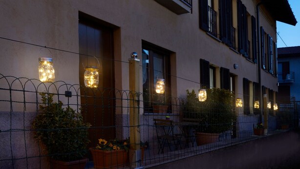 Un bâtiment doté de nombreuses fenêtres et portes, dont certaines sont équipées de luminaires. On y trouve des plantes en pot et une petite table avec des chaises à l’extérieur.