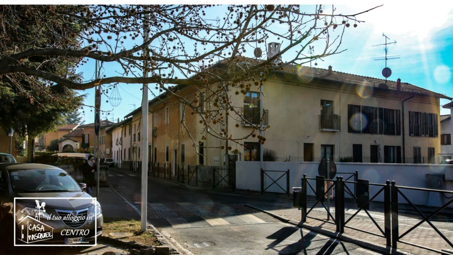 Straßenansicht eines Gebäudes mit weißer Fassade und mehreren Fenstern. Ein Auto ist am Straßenrand geparkt, und ein Fußgängerüberweg ist sichtbar.