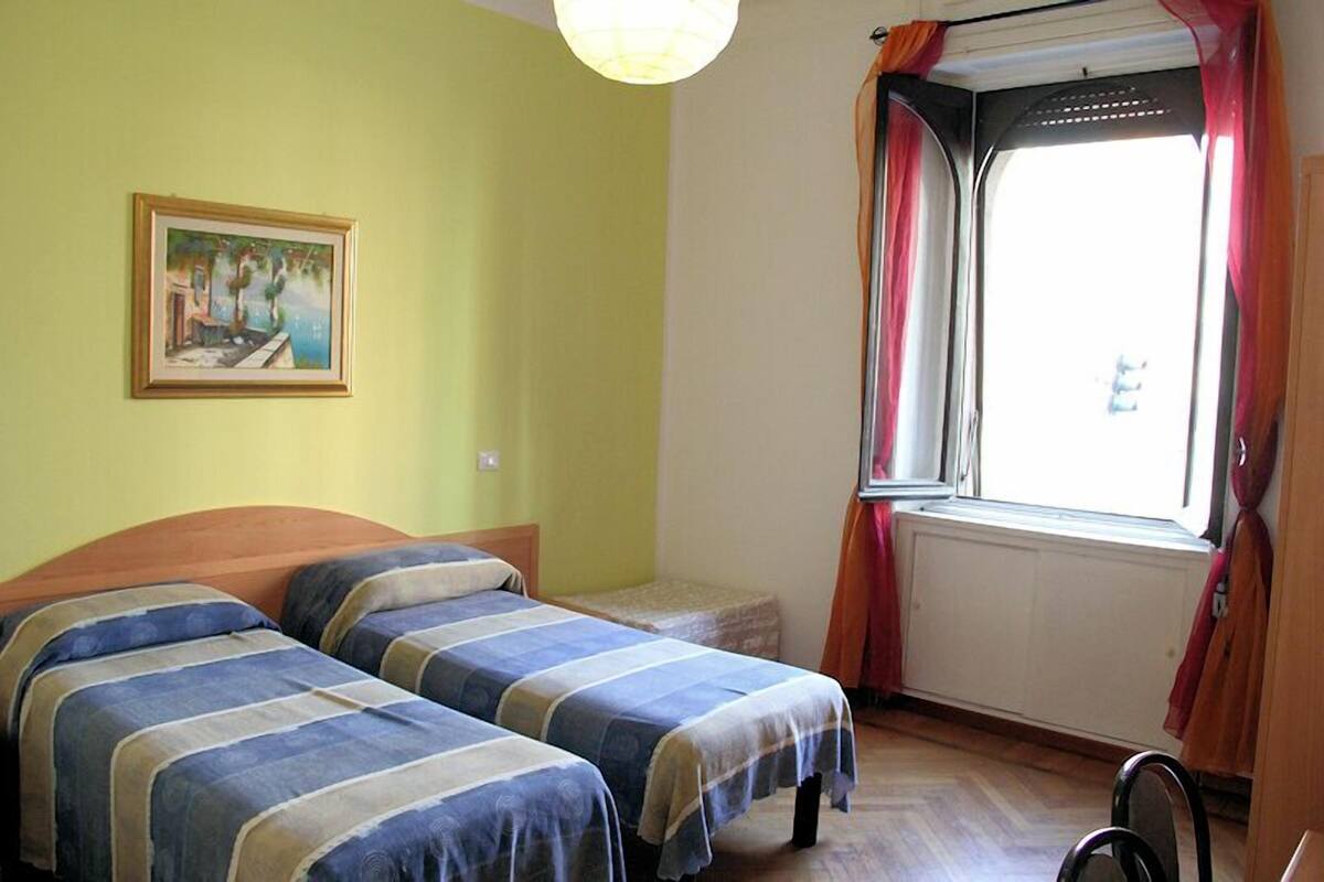 A room with two beds, a window with curtains, a radiator, and a painting on the wall.
