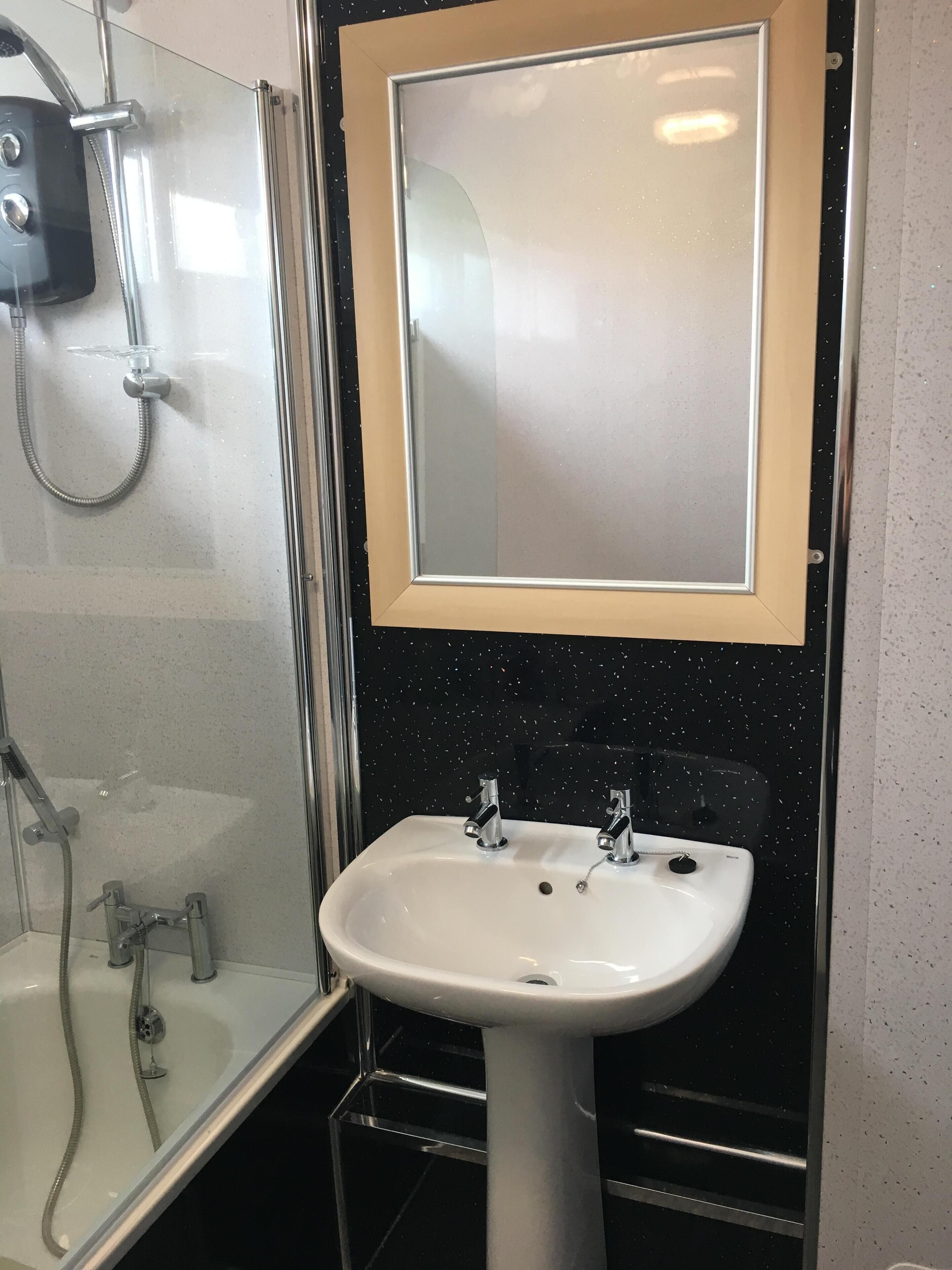 A bathroom with a glass shower enclosure, a white sink with two faucets, and a mirror with a wooden frame.