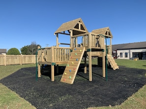 A wooden playground structure with climbing walls and a slide, set on a black rubber mat.
