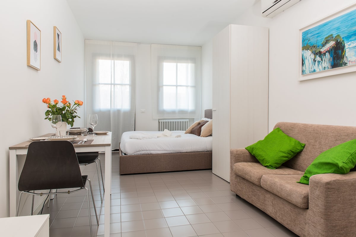 A modern, well-lit room with a bed, a sofa, a dining table, and a chair.