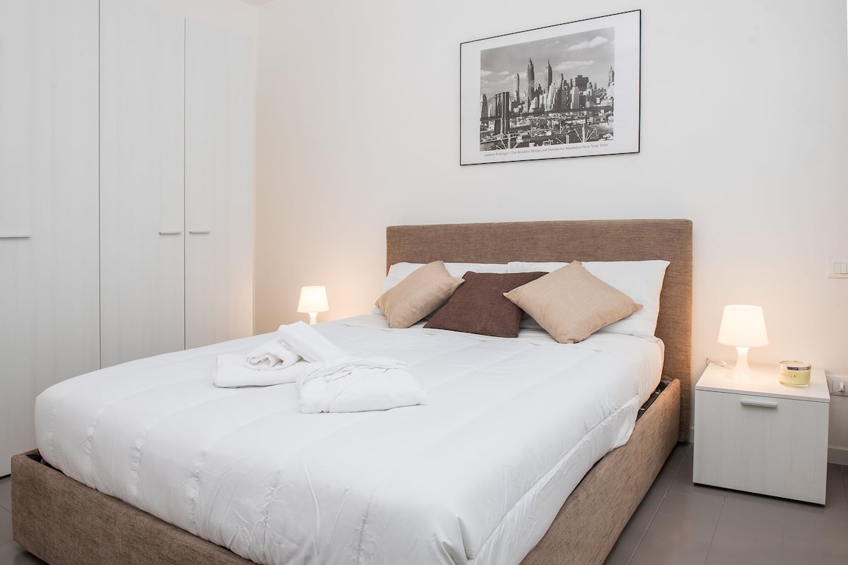 A neatly made bed with pillows, a bedside lamp, and a framed cityscape picture on the wall.