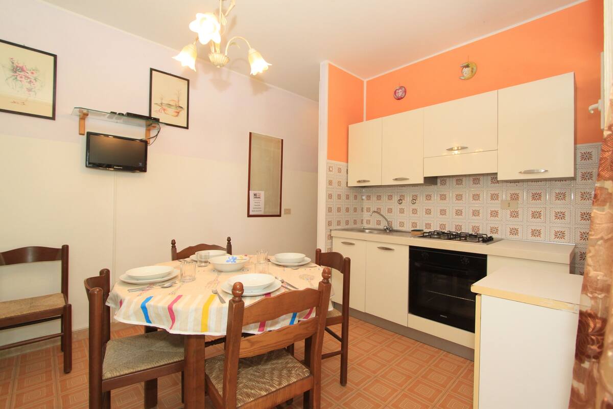 A dining area with a table set for four, wooden chairs, a television mounted on the wall, and a kitchen with white cabinets and a tiled backsplash.