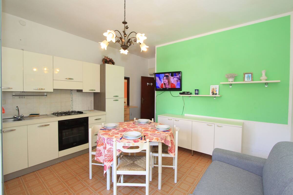 A compact kitchen with white cabinets, a dining table set for four, and a television mounted on the wall.