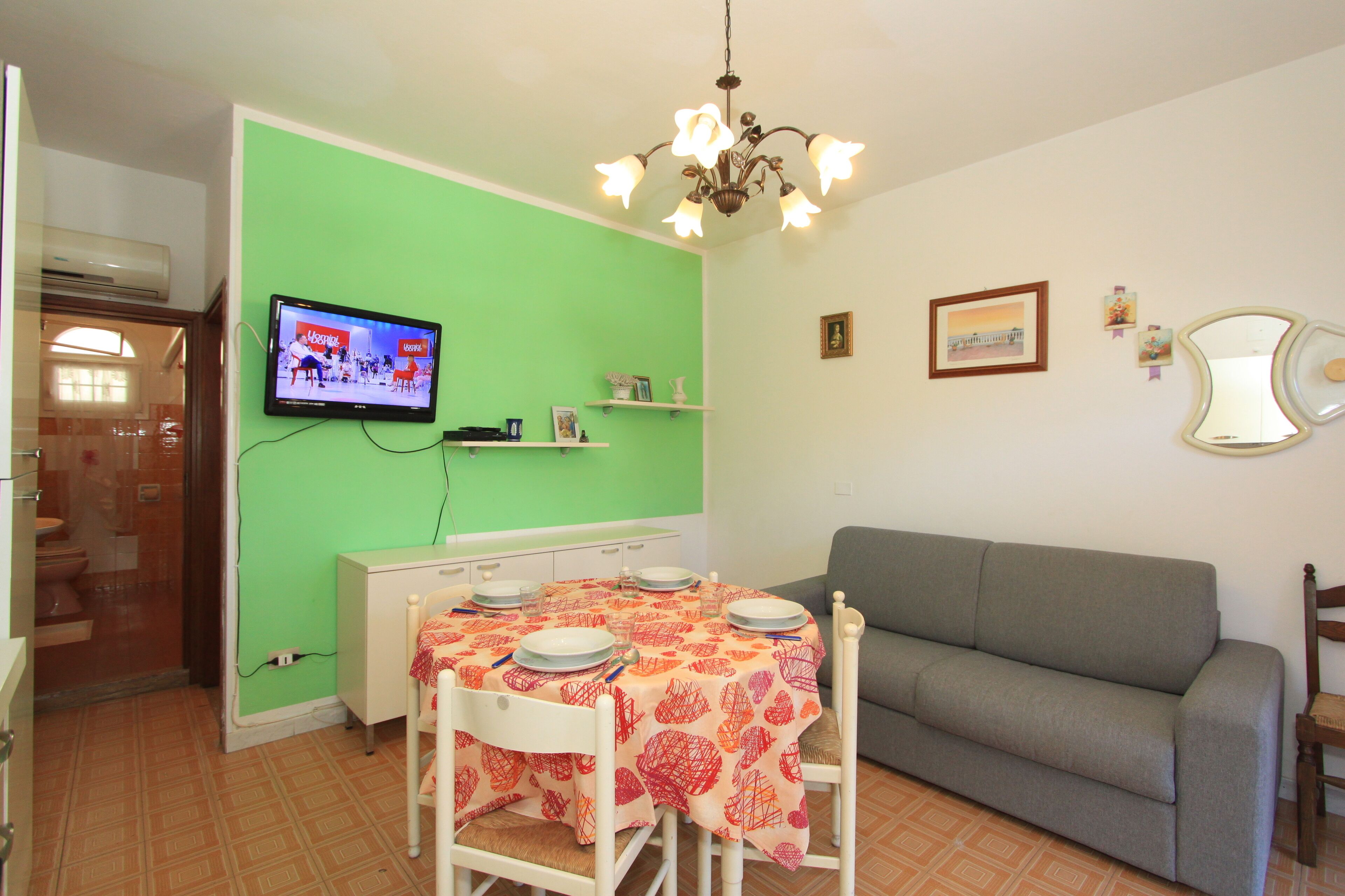A dining area with a green wall, a television mounted above the table, and a sofa in the background.