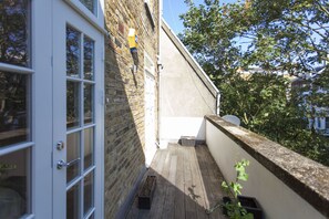 A narrow outdoor corridor with a wooden floor, a white door, and a brick wall. - Chelsea Residential Atmosphere (London)
