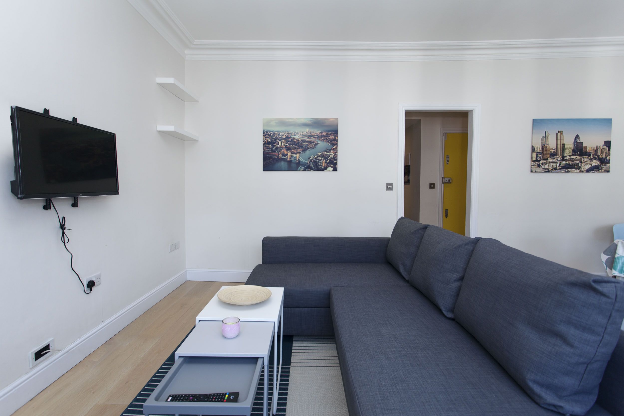 A modern living room with a grey sofa, a flat-screen TV mounted on the wall, and two cityscape pictures on the walls.