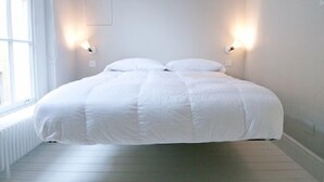Cama bem arrumada em quarto com paredes brancas e janela à esquerda.