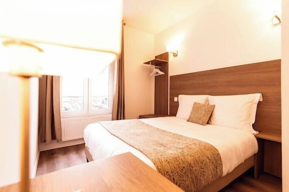 A hotel room with a wooden headboard, a bed with white linens, a bedside table, and a window with curtains.