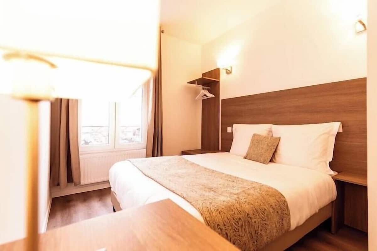Habitación de hotel con cabecera de madera, cama con sábanas blancas, mesita de noche y ventana con cortinas.