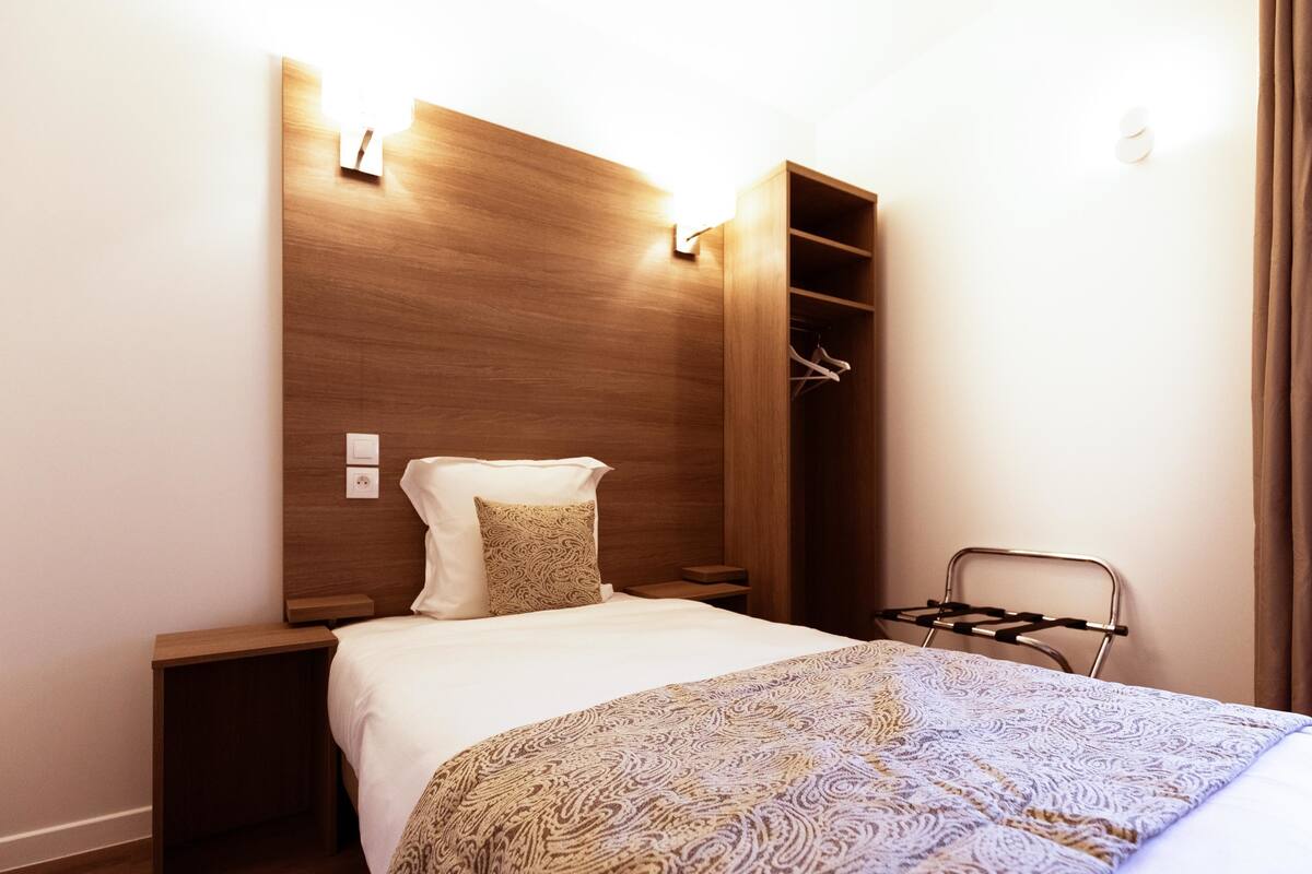 A hotel room with a wooden headboard, a bed with a patterned blanket, a hanging coat rack, and a small nightstand.