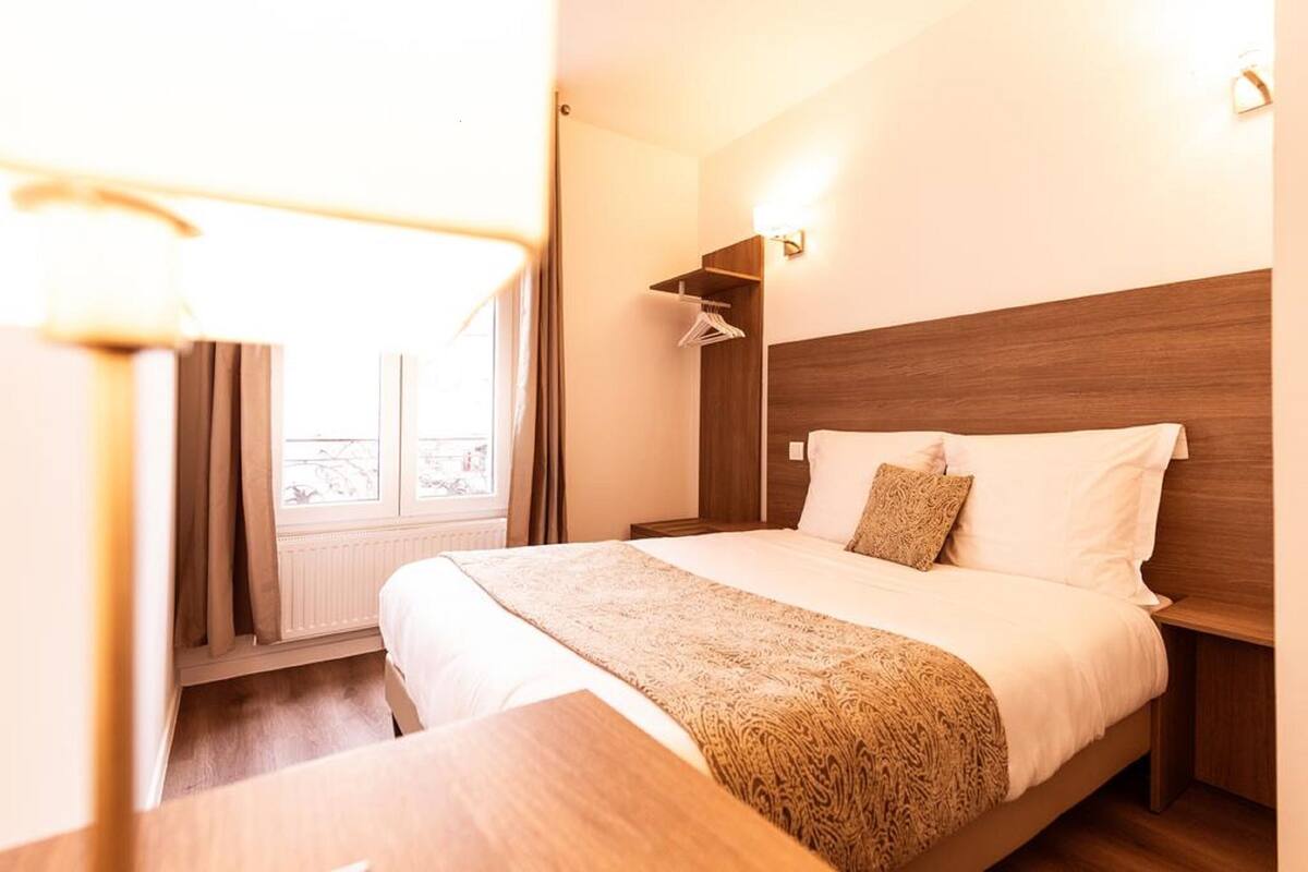 A hotel room with a bed, bedside table, lamp, window with curtains, and a wall-mounted shelf.