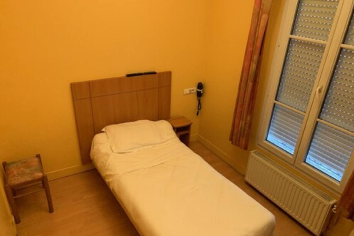 A small, single-bed room with a wooden headboard, a bedside table, a chair, and a window with blinds.
