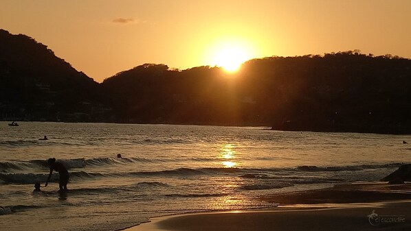 A beach scene with the sun setting over the water, people in the water, and hills in the background.