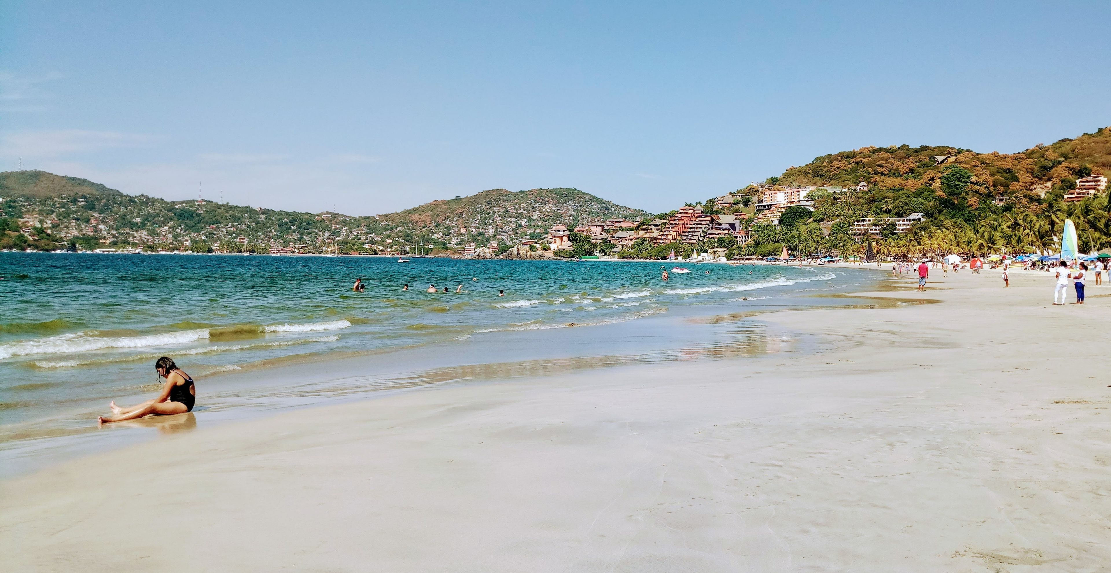 Uma praia com águas cristalinas, uma pessoa sentada na areia e edifícios ao fundo.