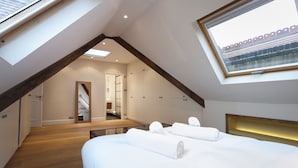 A bedroom with a skylight, wooden beams, a bed with white linens, and a view of a brick building through a window.