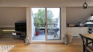 A modern living room with a large sliding glass door leading to a balcony, a wooden floor, a TV stand, and a dining area.