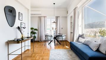 A modern living room with a blue sofa, a dining table with chairs, a large window with a city view, and a decorative mirror on the wall.