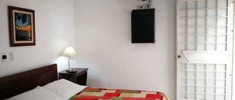 A room with a bed, a TV, a fan, a door, and a painting on the wall.