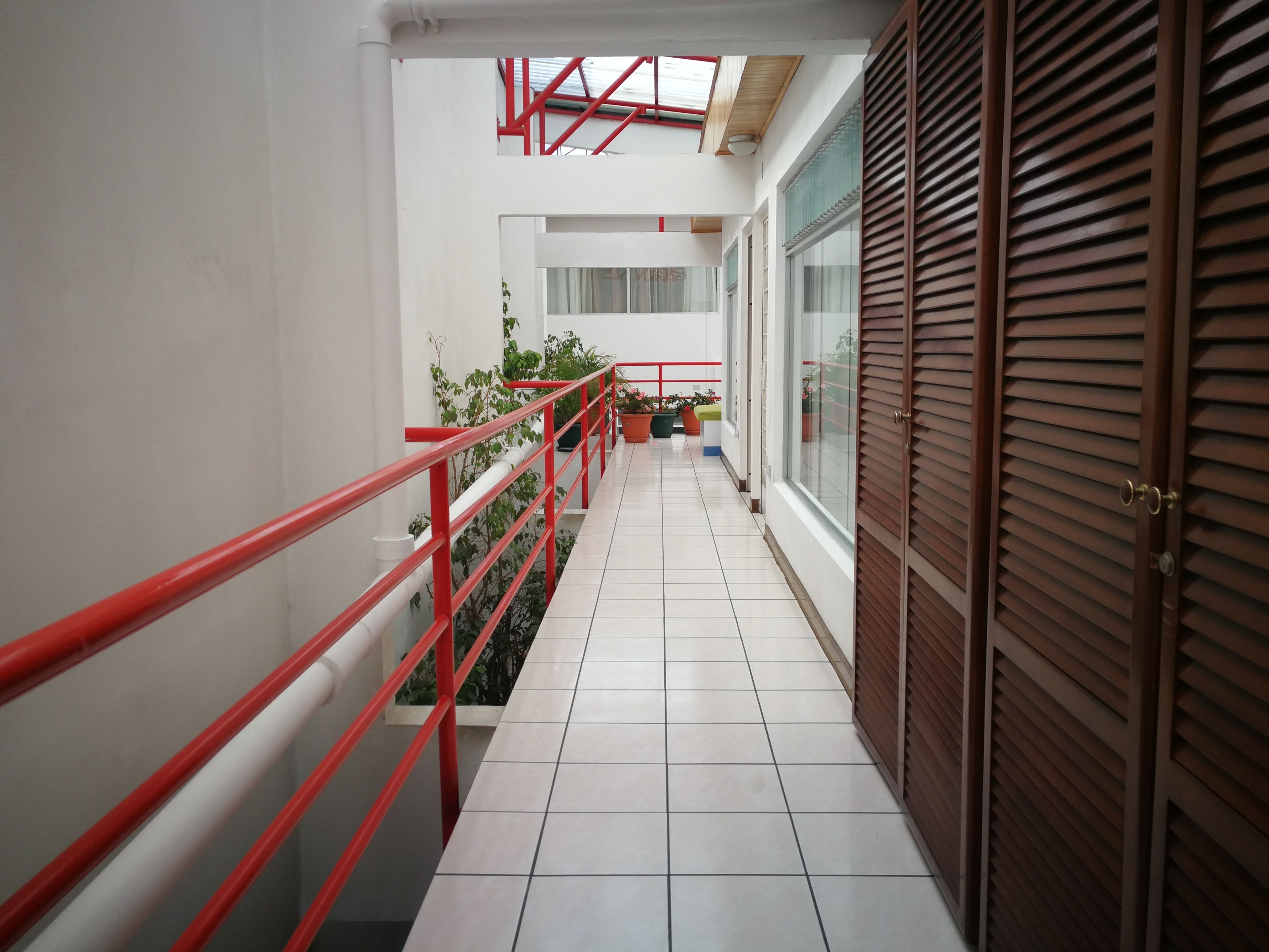 A corridor with a red railing, tiled floor, potted plants, and a wooden door.