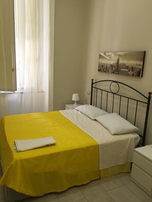 Camera da letto con un letto coperto da lenzuola gialle e bianche, un comodino bianco con una lampada e un quadro raffigurante una veduta cittadina.