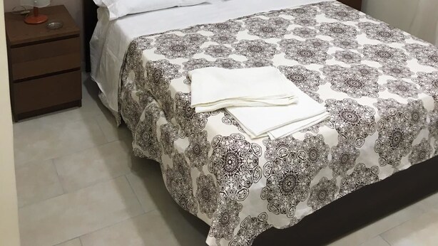 Un letto rifatto con cura, con una coperta a motivi, due cuscini bianchi e un comodino con una lampada e un piccolo vassoio.
