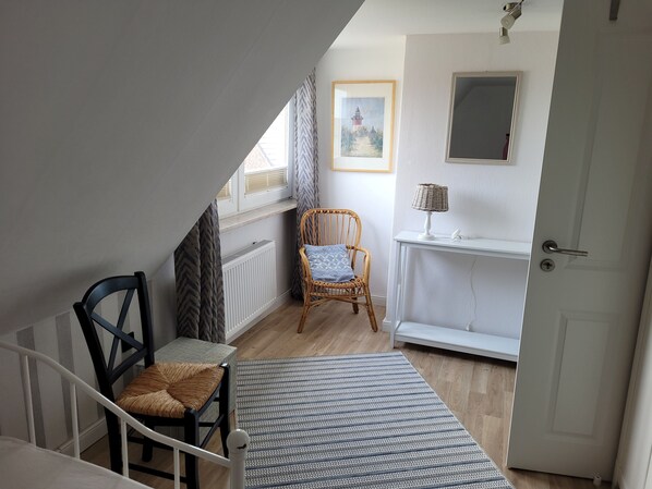 Quarto com piso de madeira, porta branca, cadeira de vime, pequena mesa, tapete estampado e um quadro na parede.