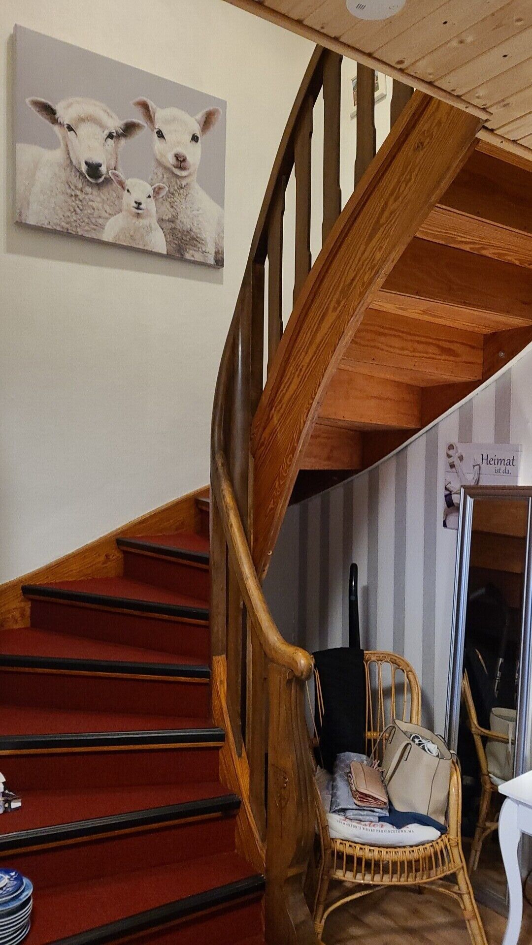 Un escalier en bois, dont les marches sont recouvertes d’une moquette rouge, mène à un palier. Une chaise en osier est placée sur le palier, et un tableau encadré représentant des moutons est accroché au mur.
