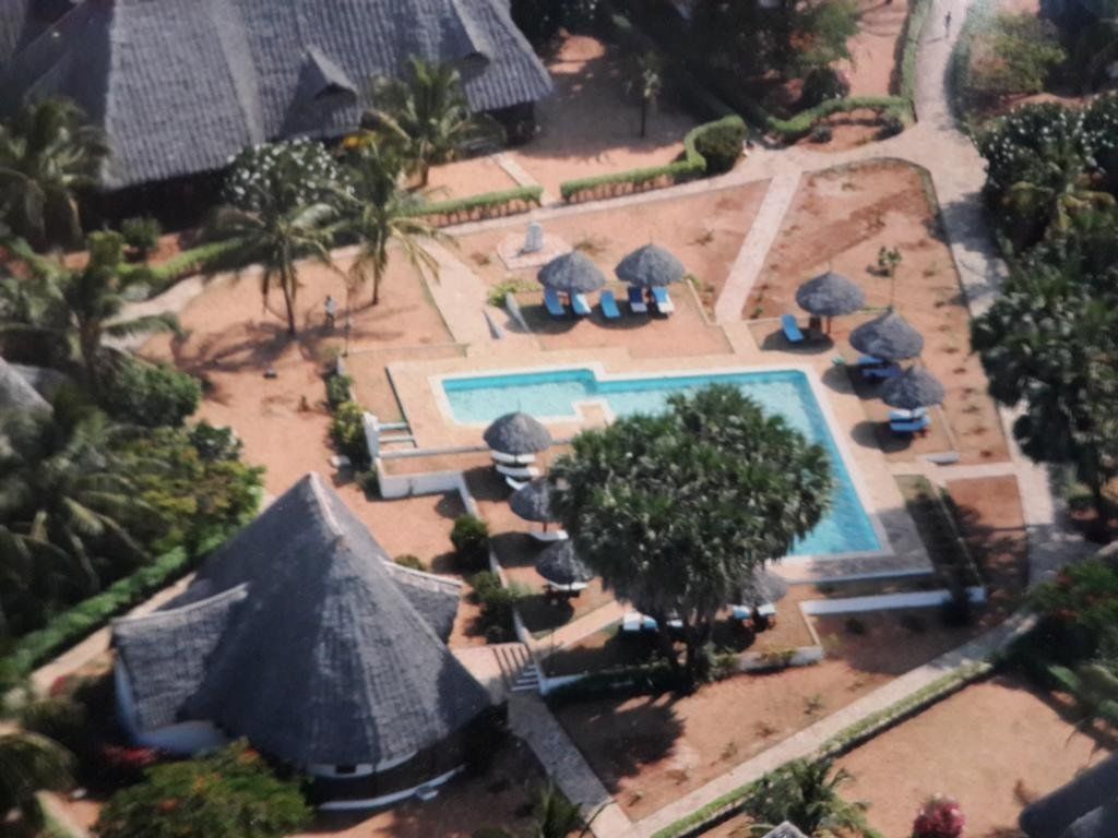Vista aérea de un resort con una piscina central, rodeada de zonas de descanso con sombrillas, palmeras y un edificio de techo de paja distintivo.