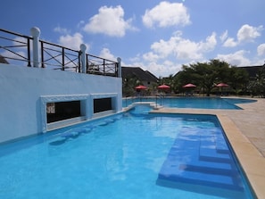 Una piscina con agua azul cristalina, rodeada por un muro blanco con diseños decorativos. Se ven escalones que bajan a la piscina, y una barandilla a la izquierda.