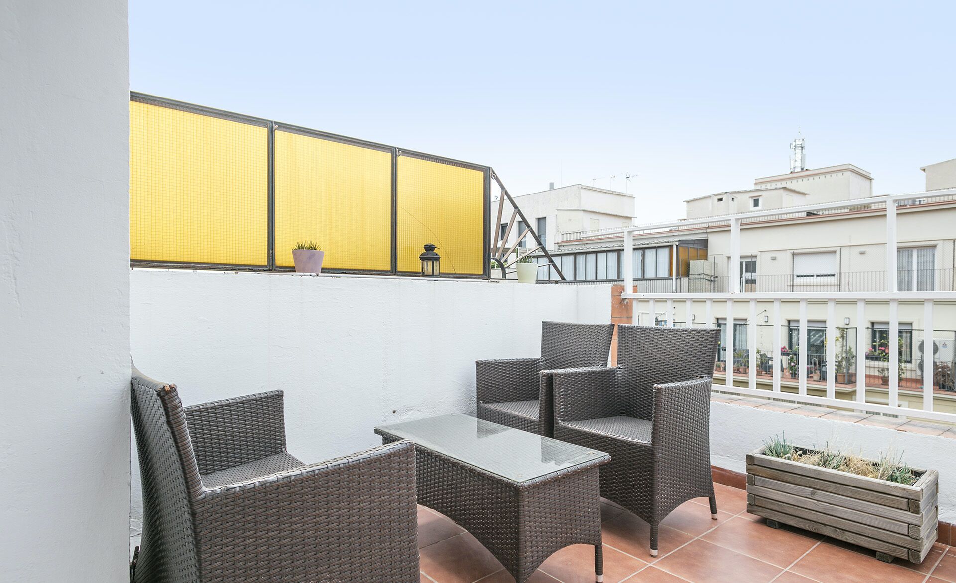 A balcony with wicker furniture and a glass table, overlooking a residential area with white buildings.