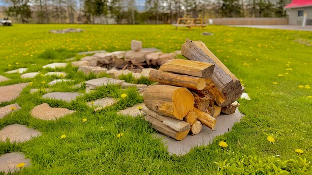 A grassy outdoor area with a stack of firewood, a stone fire pit, and a red-roofed building in the background.