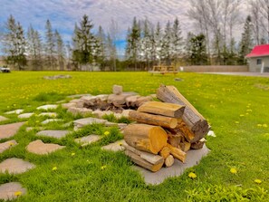 A grassy outdoor area with a stack of firewood, a stone fire pit, and a red-roofed building in the background. - Wiarton willys inn (South Bruce Peninsula)