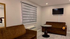 A room with a brown sofa, a small round table, a TV mounted on the wall, and a window with blinds.