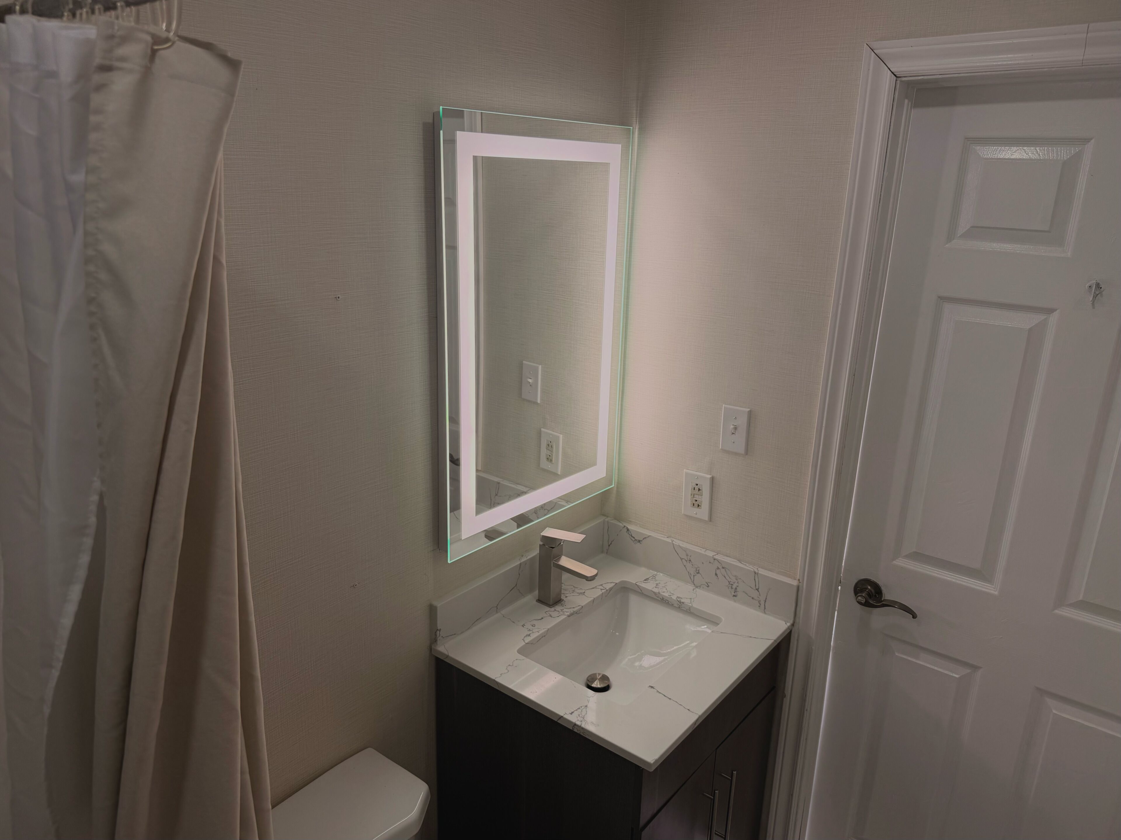 Un baño con inodoro blanco, lavabo con encimera de mármol, espejo con luz y puerta blanca.