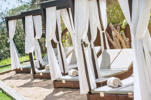 Une rangée de chaises longues d’extérieur, dotées de coussins blancs et surmontées de petits auvents, est disposée sur une surface sablonneuse, avec une végétation luxuriante en arrière-plan.