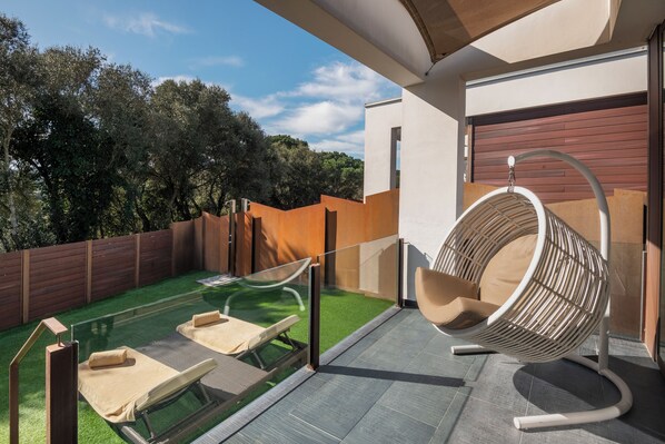 Uma área de estar moderna ao ar livre, com mesa de vidro, duas poltronas de descanso e uma cadeira suspensa.